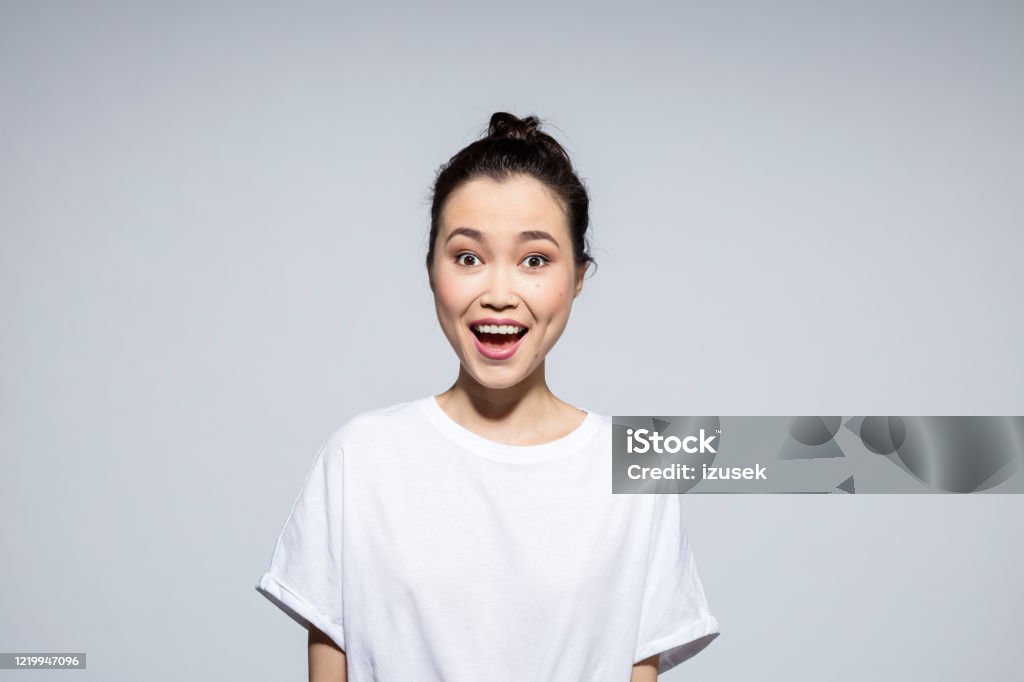 Excited young woman staring at camera Portrait of surprised asian young woman wearing white t-shirt, looking at camera with mouth open. Studio shot, grey background. Excitement Stock Photo