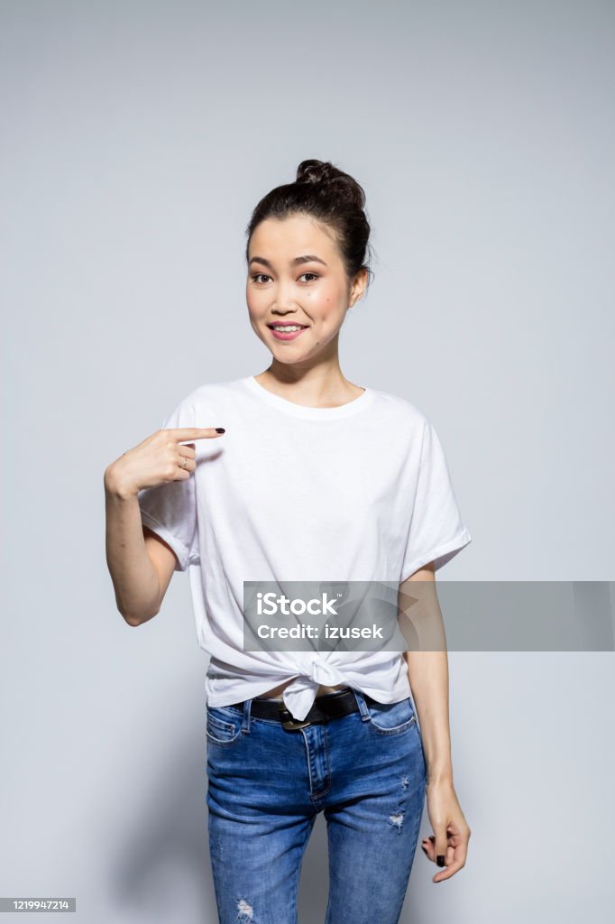Friendly asian young woman potinting at t-shirt Portrait of beautiful asian young woman wearing white t-shirt and jeans, pointing with index finger at copy space on t-shirt. Studio shot, grey background. Asian and Indian Ethnicities Stock Photo