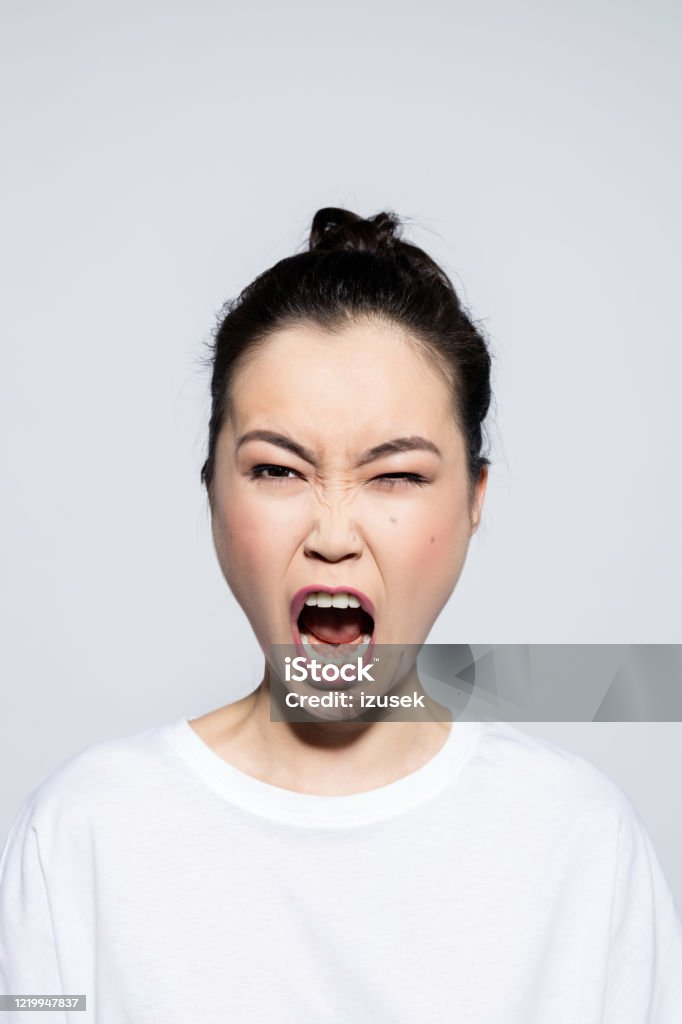 Headshot of angry asian young woman Portrait of beautiful asian young woman wearing white t-shirt, screaming at camera. Studio shot, grey background. Anger Stock Photo