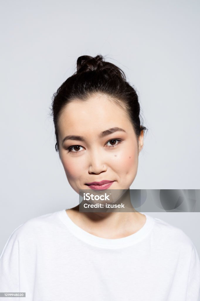 Headshot of cute asian young woman Portrait of beautiful asian young woman wearing white t-shirt, smiling at camera. Studio shot, grey background. Asian and Indian Ethnicities Stock Photo