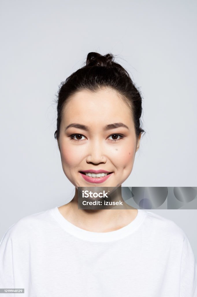 Headshot of friendly asian young woman Portrait of beautiful asian young woman wearing white t-shirt, smiling at camera. Studio shot, grey background. Front View Stock Photo