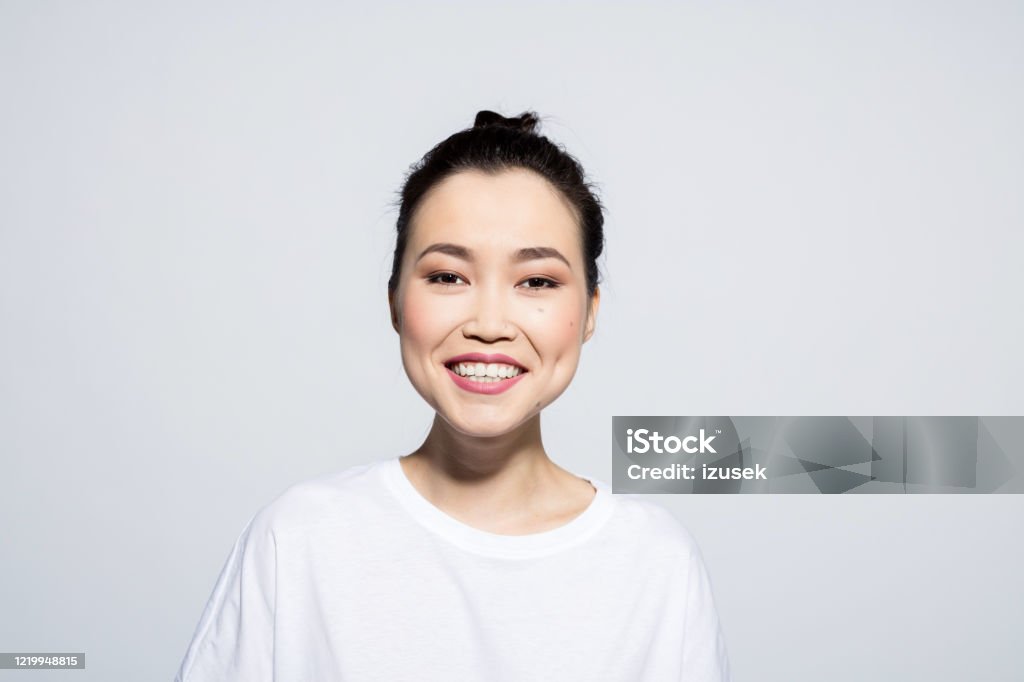 Headshot of happy asian young woman Portrait of beautiful asian young woman wearing white t-shirt, smiling at camera. Studio shot, grey background. Human Face Stock Photo
