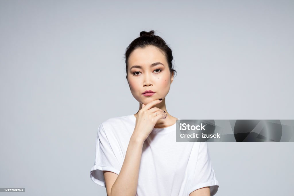 Pensive beautiful asian young woman Portrait of beautiful asian young woman wearing white t-shirt, looking at camera with hand on chin. Studio shot, grey background. Hipster Culture Stock Photo