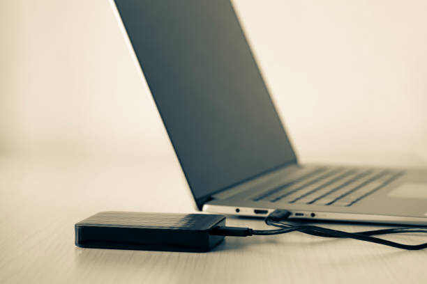istock External hard disk on wood table connected via mini usb cable to laptop, white background. Color image. 1219954462