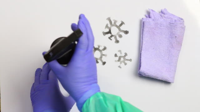 A coronavirus is painted on a white surface. A man in rubber gloves and a raincoat treats the drawing with an antiseptic and erases it with a napkin. Shot from above. Symbol of the fight against the pandemic.