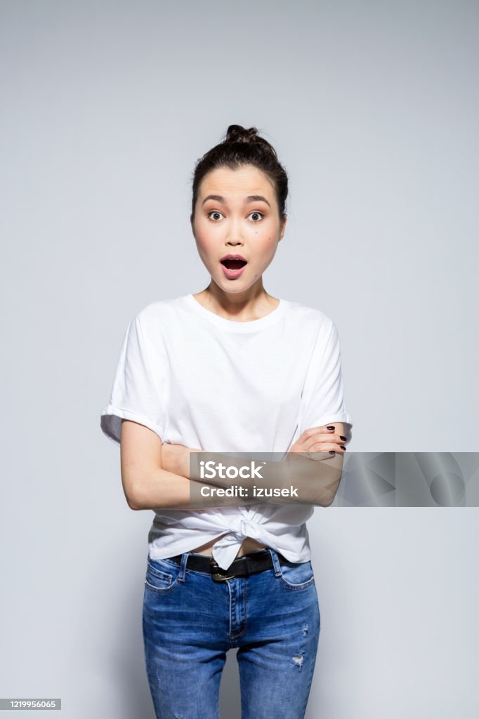 Surprised young woman staring at camera with arms crossed Portrait of beautiful asian young woman wearing white t-shirt and jeans, looking at camera with arms crossed and mouth open. Studio shot, grey background. Women Stock Photo