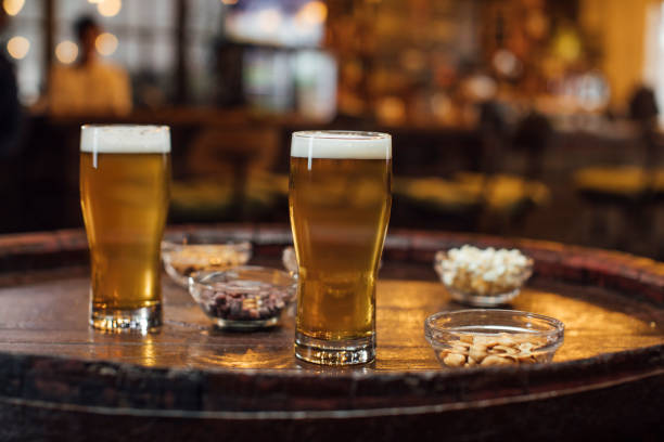 счастливый час в местном пабе: два стакана светлого пива lager и закуски на деревянном столе - beer nuts стоковые фото и изображения