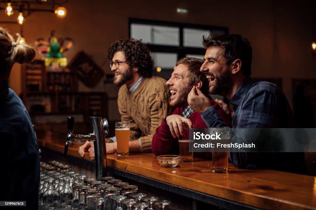 Group of Happy Young Men Drinking Beer and Watching a Soccer Game at the Pub Group of friends drinking beer and snacking while watching football at the bar. Pub Stock Photo