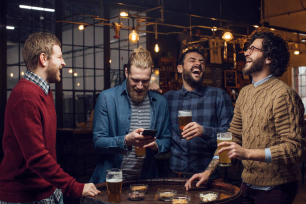 Group of Men Having a Laugh at the Pub While Drinking Beer and Watching a Football Game on the Mobile Phone Four male friends having fun at the bar together by watching football and drinking beer. friends in bar with phones stock pictures, royalty-free photos & images