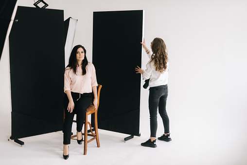 Young woman photographer is preparing backdrop in studio for photo shoot. Female model sits on chair and looks into the camera, waiting for work to begin. Concept of backstage job in photo studio.