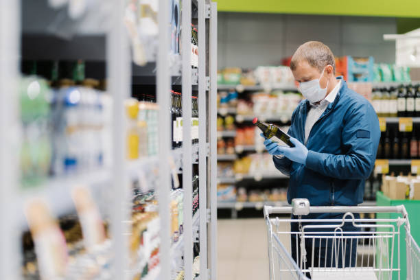 zdjęcie człowieka w ochronnej masce medycznej i gumowych rękawicach zapobiegających koronawirusowi, pozuje w supermarkecie, trzyma szklaną butelkę napoju alkoholowego, czyta etykietę, robi zakupy w czasie kwarantanny - isolated on white bottle alcohol alcoholism zdjęcia i obrazy z banku zdjęć