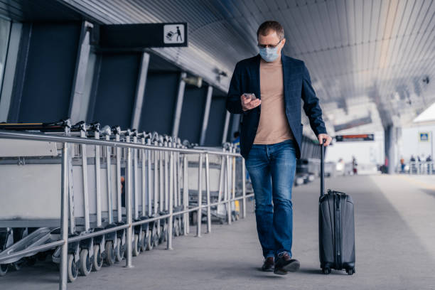 Male passenger walks in airport, holds suitcase, checks newsfeed on smartphone, wears disposable medical mask during pandemic coronavirus, tries to be safe during disease virus. Respiratory symptoms pasajero masculino camina en el aeropuerto, sostiene la maleta, revisa la noticiasde fiela en el teléfono inteligente, usa máscara médica desechable durante el coronavirus pandémico, intenta estar a salvo durante el virus de la enfermedad. síntomas re