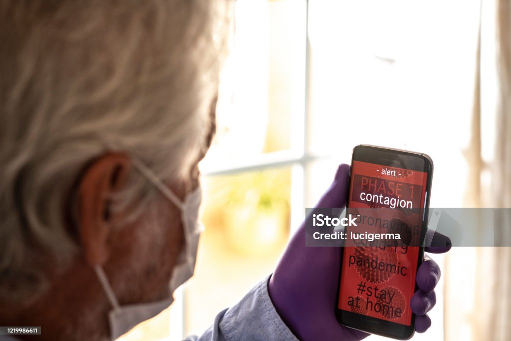 Senior man wearing facial mask and gloves receives alert on mobile phone app to stay at home avoiding contagion by coronavirus Covid-19. Active Seniors Stock Photo