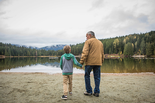 Grandfather walking with his grandson on sandy beach toward calm lake on overcast, autumn\tday.