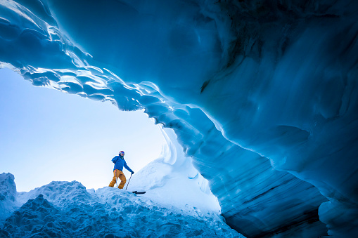 Male skier in 30’s wearing yellow pants and blue winter jacket looking into ice cave in Whistler backcountry.