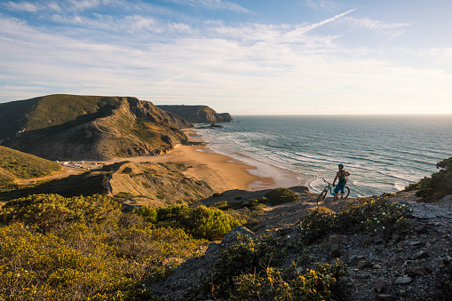 Fit man standing with his mountain bike on top of cliff looking at ocean.