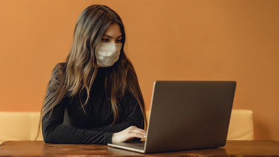 Portrait of adult woman with medical face mask  on yellow background. Concept with copy space. She is working with modern gray laptop