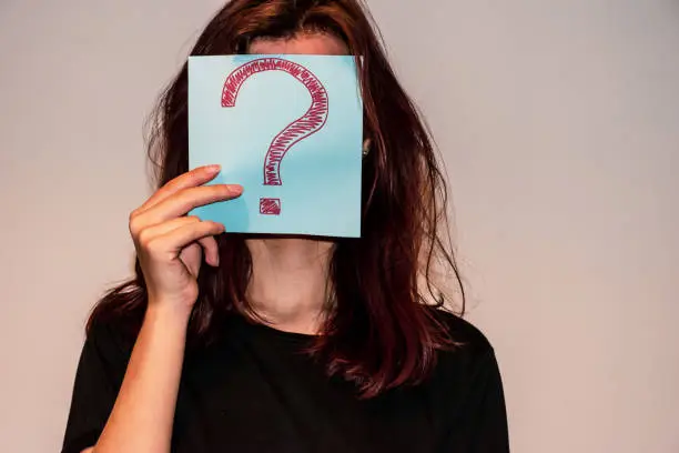 Photo of A close-up shot of an unrecognizable young Caucasian redhead woman hiding her face behind a light blue paper sheet with a hand-drawn question mark