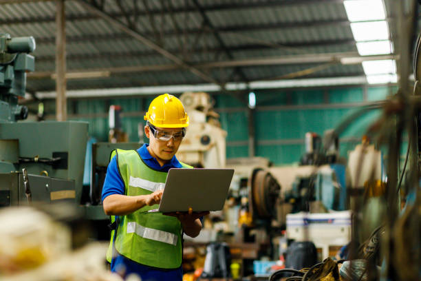 istock Men industrial engineer in uniform and wearing a Yellow helmet while standing in a heavy industrial factory behind he looking New Project while Using Laptop. 1220021458