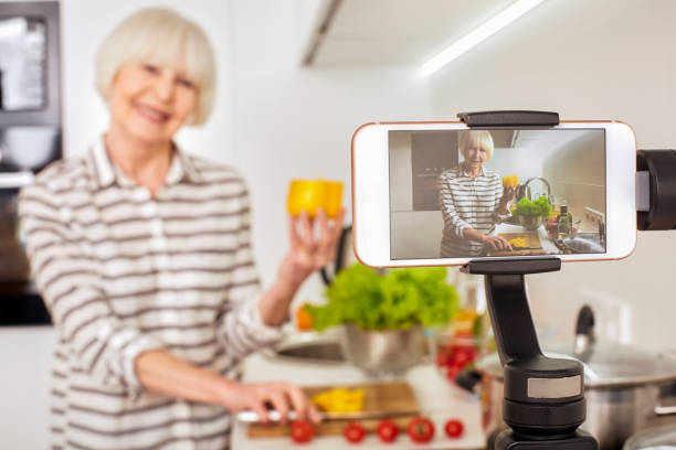 Blurred background isolated on kitchen light interior.Aged female old woman record broadcast live video online cook healthy fitness diet summer food. Lady vlogger blogging has followers youtube chanel