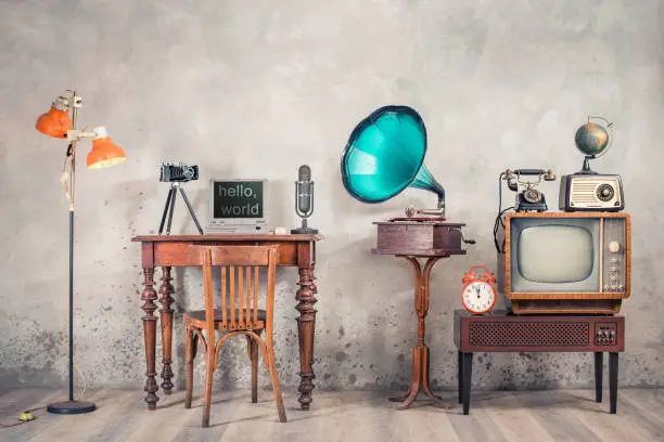 Photo of Retro TV, old radio, telephone, alarm clock, globe, gramophone, classic microphone, outdated laptop and camera on wooden table, floor lamp front concrete wall background. Vintage style filtered photo