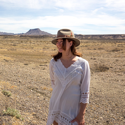 Woman with a hat and long hair standing in desert with sand and rock formations behind her.