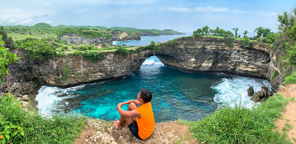 Tourist man sitting on the cliff looking to the an incredibly beautiful beach with beautiful rocks. Aerial & panorama view of Broken Beach ,Angel's BillaBong Beach, Nusa Penida island, Bali, Indonesia.