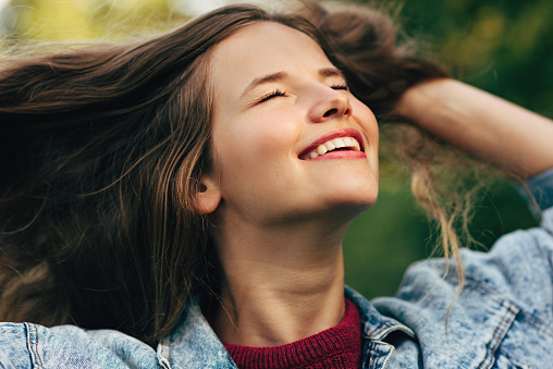 Close-up portrait of beautiful young woman smiling broadly with healthy toothy smile, with closed eyes, wearing denim jacket, posing on nature background in the park. People, travel, lifestyle