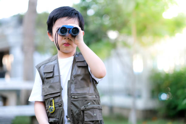 istock Curious little young Asian boy looking through a binoculars in the garden. Outdoors activity. Feeling fun. Boy wear green outdoors vest which decorate with compass 1220071241