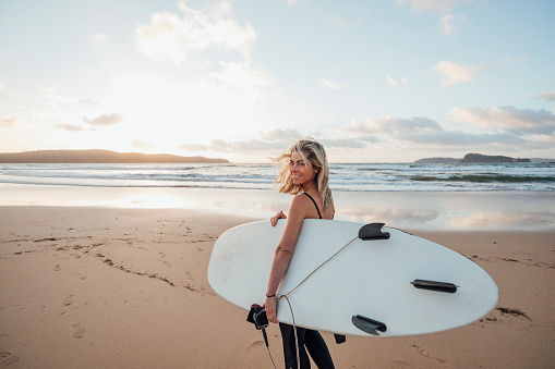 A side view shot of a woman wearing a wetsuit, she is standing on the beach and holding a surfboard, she is looking at the camera.