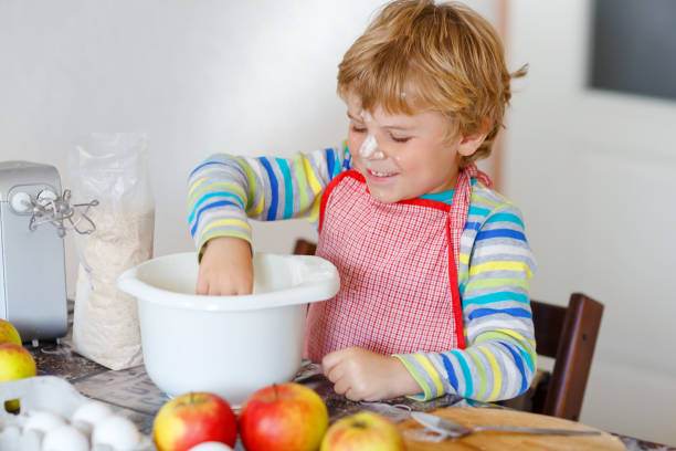 cute little happy blond preschool kid boy baking apple cake and muffins in domestic kitchen. funny lovely healthy child having fun with working with mixer, flour, eggs, fruits. little helper indoors - cake making mixing eggs imagens e fotografias de stock