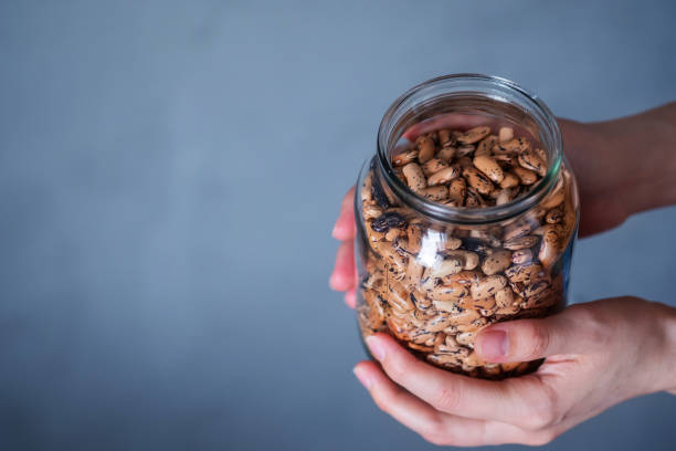 istock Hands holding a large glass jar with raw beans on a gray background on the left - an empty place for text. Offering veggie food 1220097639
