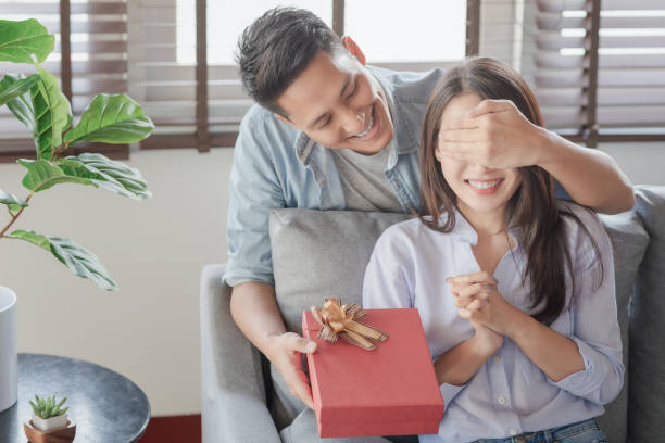 homem bonito está dando a sua namorada uma caixa de presente vermelho para o aniversário surpreendente na sala de estar - family loving anniversary gift - fotografias e filmes do acervo
