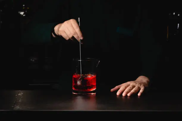 Closeup shot of bartender hand stirring a negroni cocktail. Glass of drink on counter with orange peal