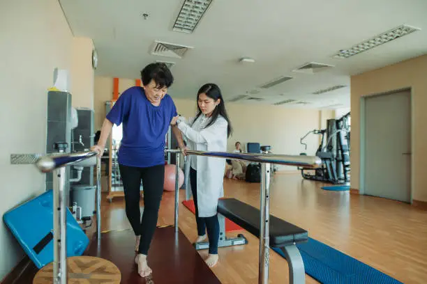 Photo of Asian chinese senior women holding parallel bars and walking while physical therapists helping at side in a rehab centre at hospital