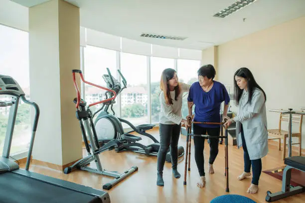 Photo of Asian chinese senior women holding parallel bars and walking while physical therapists helping at side in a rehab centre at hospital