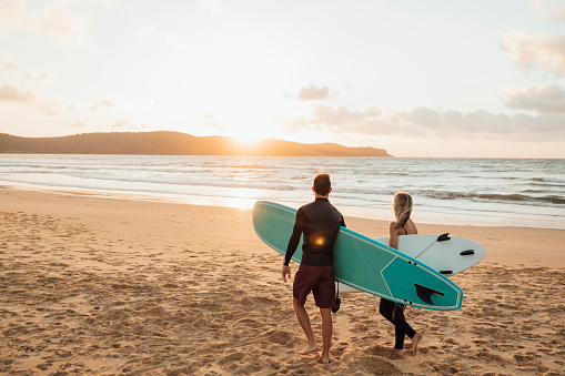 A rear-view shot of a young man and woman holding a surfing board and walking on a beach in Sydney, Australia.