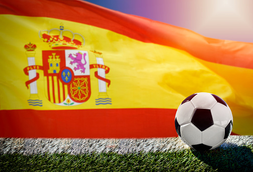Soccer or football on green grass with Spanish waving flag in background