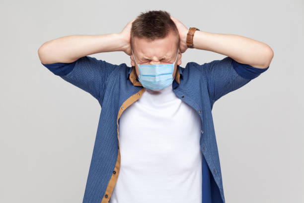Headache. Portrait of young man in casual style with surgical medical mask standing and holding his painful head. medical and health care concept. Headache. Portrait of young man in casual style with surgical medical mask standing and holding his painful head. medical and health care concept. indoor studio shot, isolated on gray background. Phobia stock pictures, royalty-free photos & images