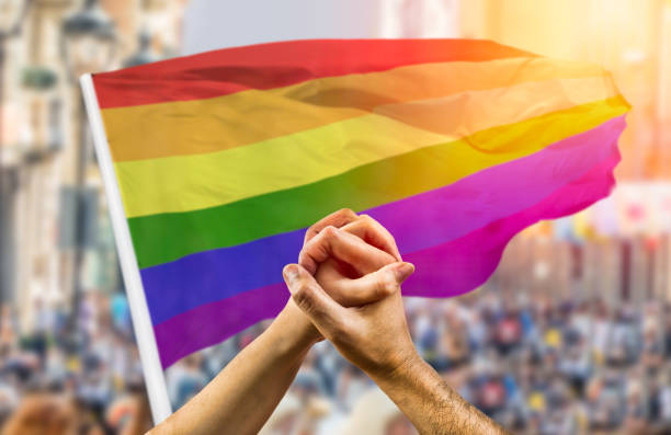 paar hält hände und winkt vor einer regenbogenfahne - homosexual gay man parade flag stock-fotos und bilder