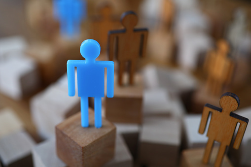 Close-up view of wooden human models placed on blocks. Blue plastic person on centre stage. Stand out from the crowd and leadership and uniqueness concept