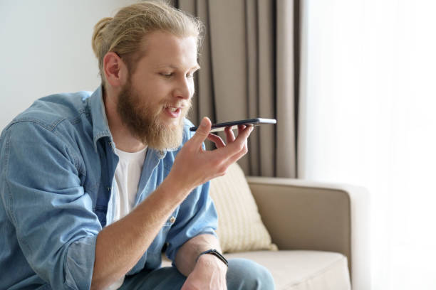 Young man holding smart phone speak on speakerphone, using virtual digital voice recognition assistant search, record audio message on smartphone at home. Mobile app ai digital tech assistance concept