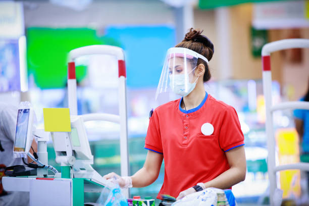 female supermarket cashier in medical protective mask and face shield working at supermarket - asian ethnicity shopping mall supermarket store imagens e fotografias de stock