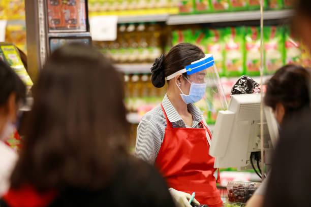 female supermarket cashier in medical protective mask and face shield working at supermarket - asian ethnicity shopping mall supermarket store imagens e fotografias de stock