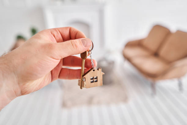 istock Men hand holding key with house shaped keychain. Modern light lobby interior. Mortgage concept. Real estate, moving home or renting property. 1220142209