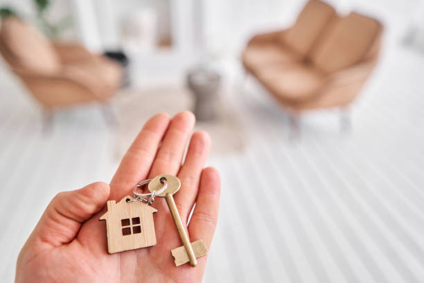 istock Men hand holding key with house shaped keychain. Modern light lobby interior. Mortgage concept. Real estate, moving home or renting property. 1220142262