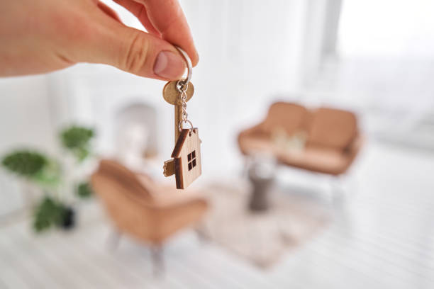 istock Men hand holding key with house shaped keychain. Modern light lobby interior. Mortgage concept. Real estate, moving home or renting property. 1220142352