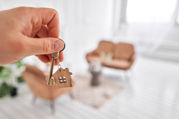 istock Men hand holding key with house shaped keychain. Modern light lobby interior. Mortgage concept. Real estate, moving home or renting property. 1220142361