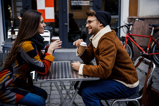 Gladsome young man and woman smiling to each other on the cafe terrace and drinking coffee from paper cups