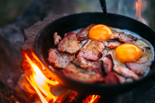Fried eggs with bacon in a pan in the forest. Food at the camp. Scrambled eggs with bacon on fire. Lots of seasonings on fried eggs. Picnic. Cooking on an open fire